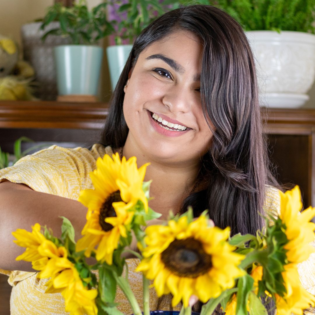 Image of a brunette wearing a yellow shirt, smiling into the camera as she holds a vase of sunflowers.
