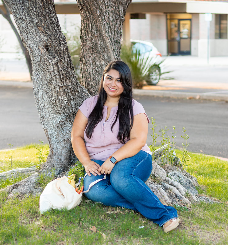 Woman sitting in the grass, leaning against a tree and smiling off into the distance. You see a canvas bag filled with veggies and sunflowers perched next to her.